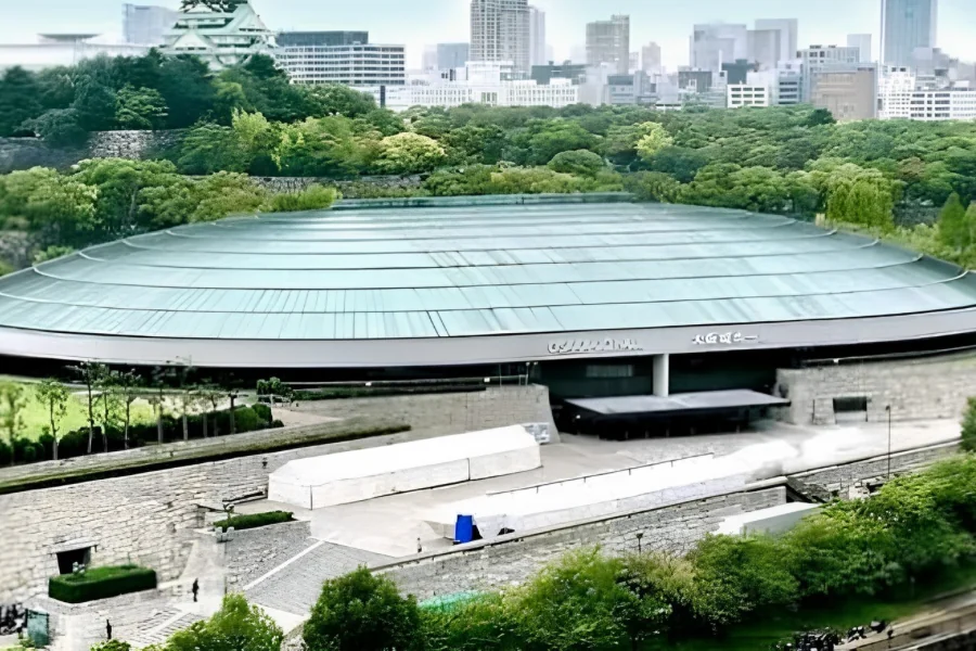 大阪城公園の緑に囲まれた大阪城ホールの外観。円形の淡い緑色の屋根の奥に大阪城の天守閣とビル群が見える
