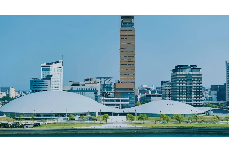 あなぶきアリーナ香川の外観。瀬戸内海ごしに見た建物で、白いなだらかなドーム状の屋根が2つ並び、背後に高松シンボルタワーとサンポート高松のビル群が立っている