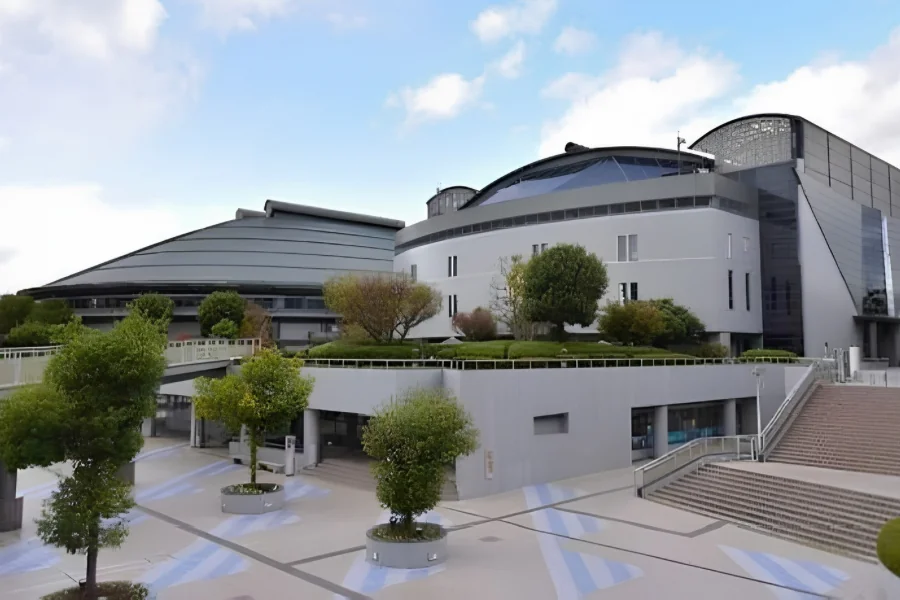 広島グリーンアリーナの外観。手前の広場に円形の植え込みと並木があり、奥に円筒形の白い建物と、左側に大アリーナの湾曲した屋根が見える