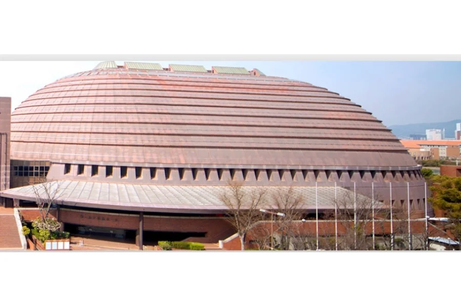 神戸ワールド記念ホールの外観。楕円形の赤茶色のドーム屋根を持つ建物が街並みの中に建ち、低層部に三角形の開口が並んでいる