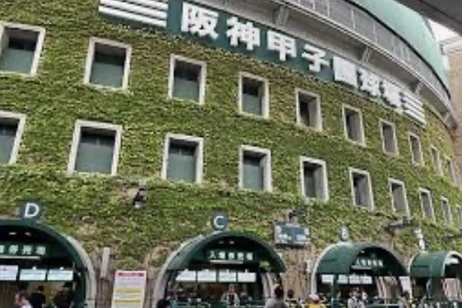 ツタに覆われた阪神甲子園球場の外壁。緑の壁面に球場名の看板が掲げられ、アーチ型の入場口の前を人が歩いている