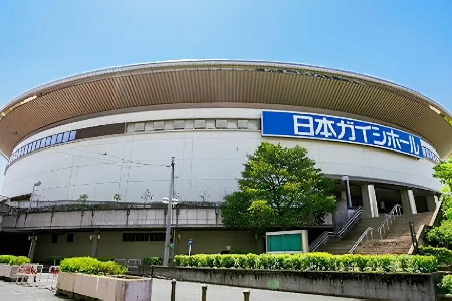 日本ガイシホールの外観。白い円形の建物に茶色い庇のような屋根がかかり、青い看板に「日本ガイシホール」と白抜きで書かれている。手前は植え込みと階段