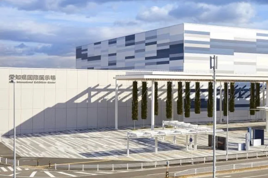 愛知県国際展示場の外観。白い壁に「愛知県国際展示場」の館名表示が入り、奥に市松模様の壁面をもつ建物が立つ。手前は柱の並ぶ屋根つきの広場